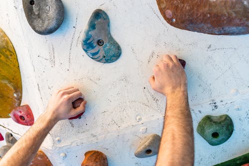 Unrecognizable sportive male mountaineer climbing artificial steep white wall while grabbing colorful climbing holds during bouldering exercise in professional gym