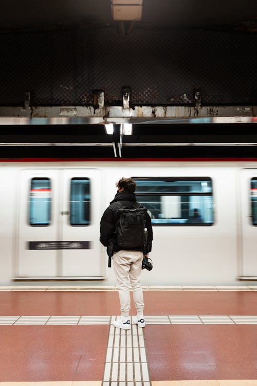 Full body back view of anonymous male standing on platform of subway station near moving train while waiting for transport on blurred background