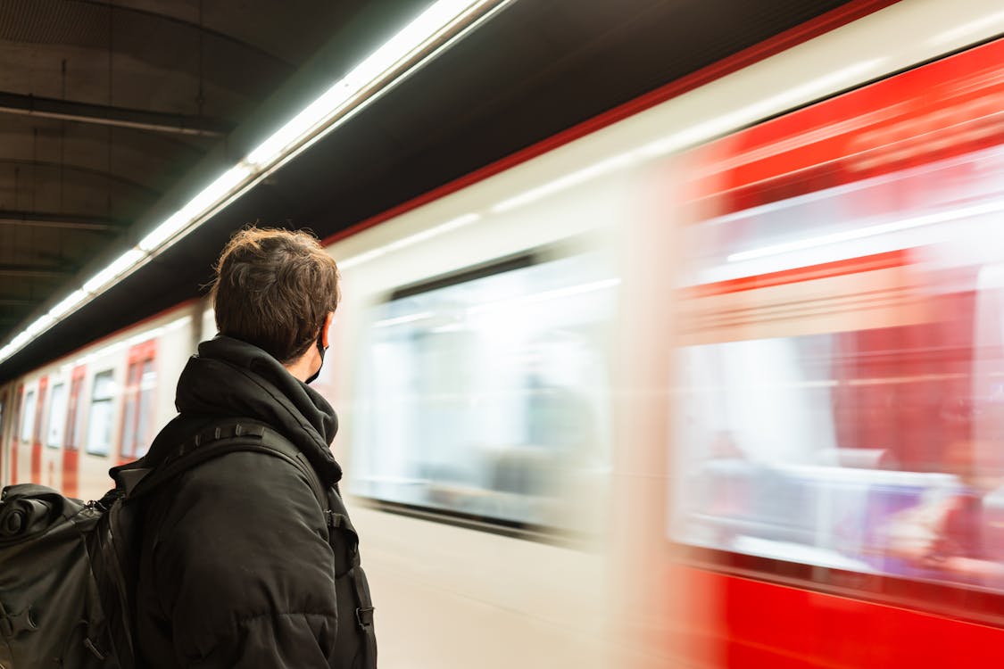 Side view of faceless male passenger in protective mask wearing warm outerwear standing on platform of metro station near train