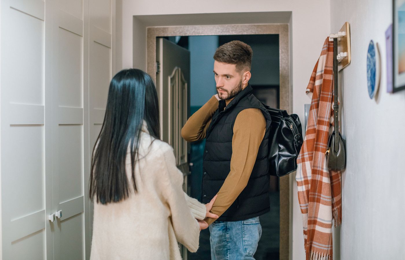 A couple holds hands in a hallway as a man prepares to leave with a backpack.