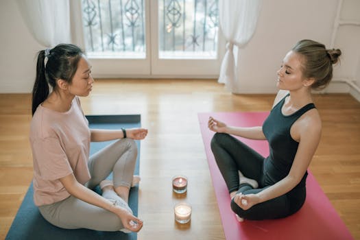 Two women practicing meditation indoors, sitting cross-legged on yoga mats beside lit candles.