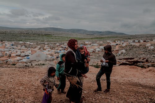 Poor ethnic woman with toddler in arms and kids near settlement surrounded with mountains under cloudy gloomy sky