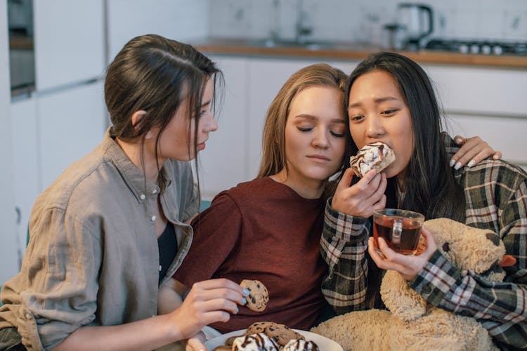 Friends Supporting A Sad Young Woman Sitting On A Sofa And Eating Pastry And Drinking Tea