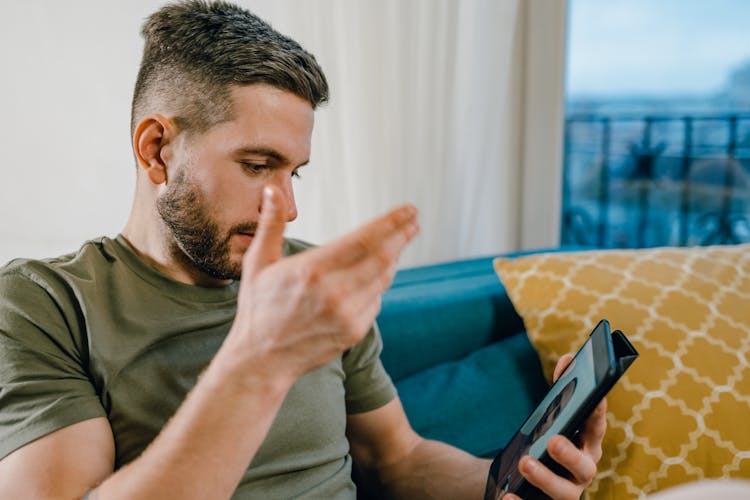 A Bearded Man In Green Shirt Sitting On The Couch While Talking To His Friend On Tablet