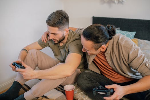 Two male friends enjoying a relaxed gaming session indoors, sitting closely on a bed.
