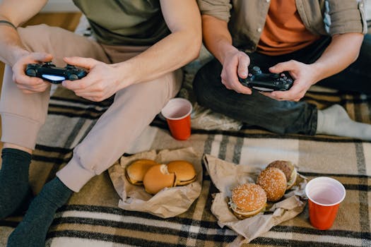 Two friends play video games together while enjoying burgers and drinks indoors.