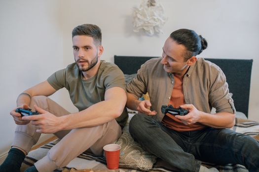 Two adult men sitting on a bed and playing video games with controllers, enjoying leisure time.