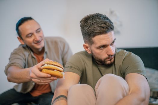 A man humorously offers a burger to his friend who seems uninterested or upset.