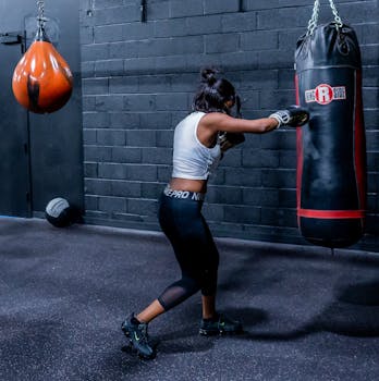 A woman in activewear practicing boxing with a punching bag in a gym setting, emphasizing strength and fitness.