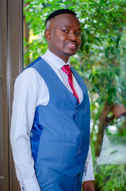 Cheerful African American male in festive blue suit and white shirt smiling and looking at camera against green trees in daylight