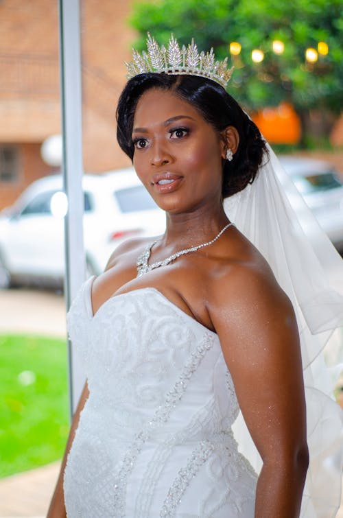Pensive African American bride with dark hair in white wedding dress and veil standing on street and looking away in daylight