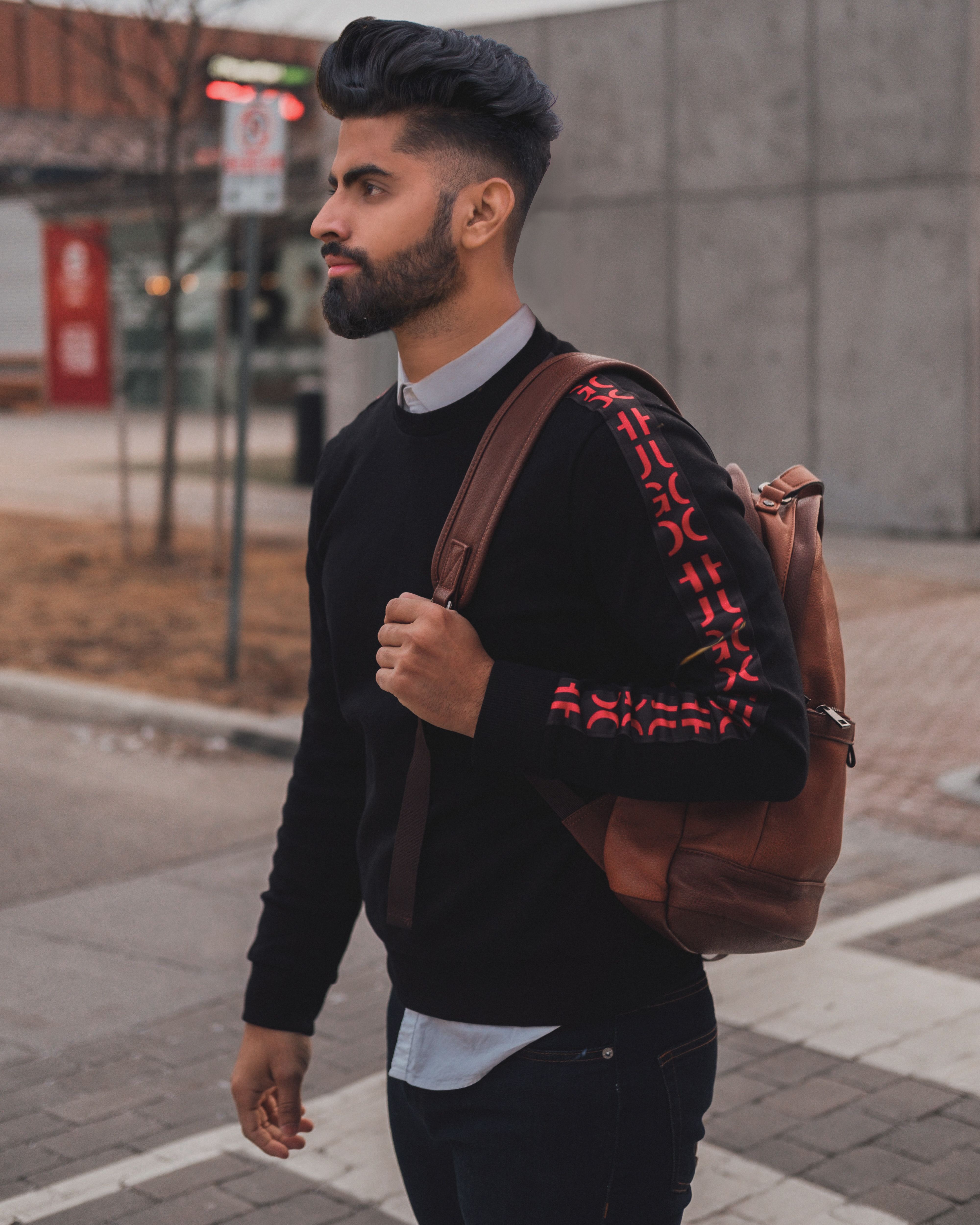 A fashionable adult male walking outdoors with a stylish outfit and backpack in a modern urban environment.