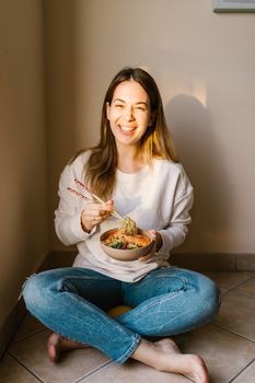 A smiling woman sits on the floor enjoying a bowl of Asian cuisine with chopsticks.
