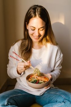 Happy woman in white sweater savoring a bowl of delicious seafood noodles with chopsticks.