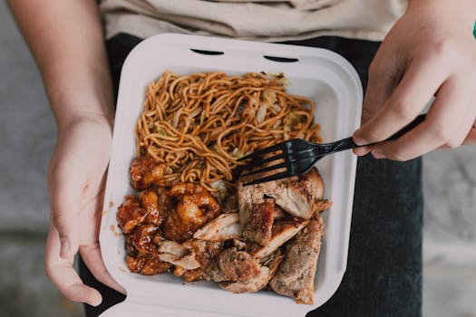 Close-up of takeout box with noodles, chicken, and pork, held in hands.