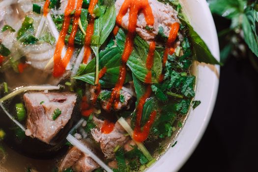 Close-up of Vietnamese pho with fresh herbs, beef, noodles, and Sriracha sauce.