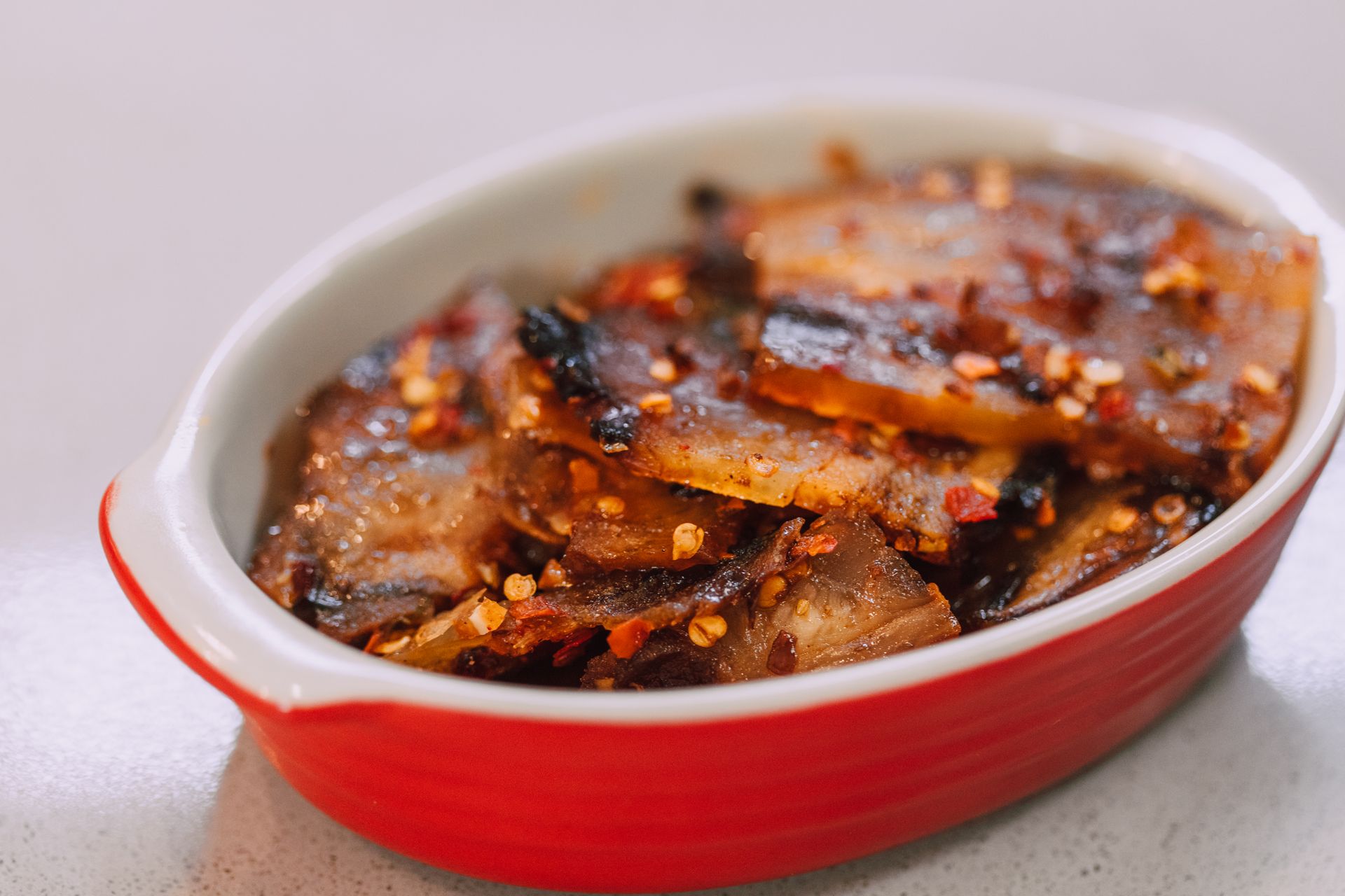 Delicious grilled pork belly seasoned with chili flakes in a red ceramic dish on a white table.