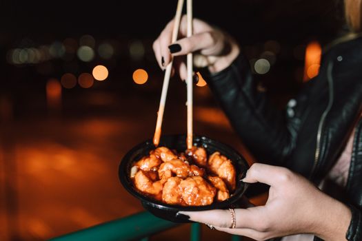 A person holds chopsticks above a bowl of saucy Asian food with bokeh night lights in the background.