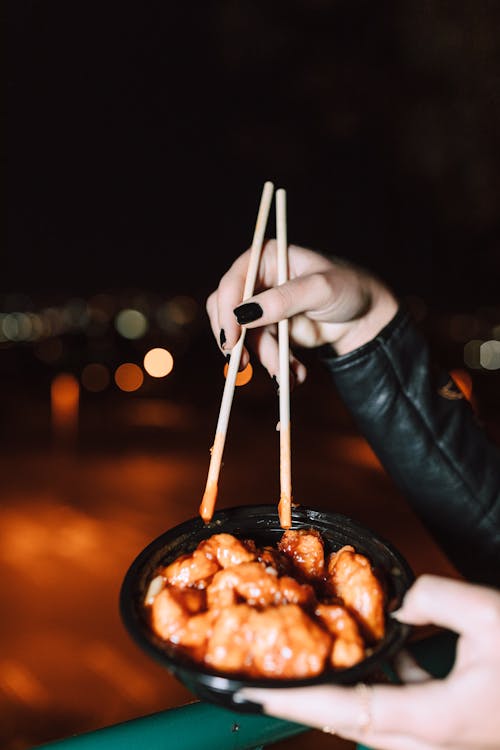 Free A hand with chopsticks hovers over a bowl of delicious Asian food, set against a blurred nighttime background. Stock Photo