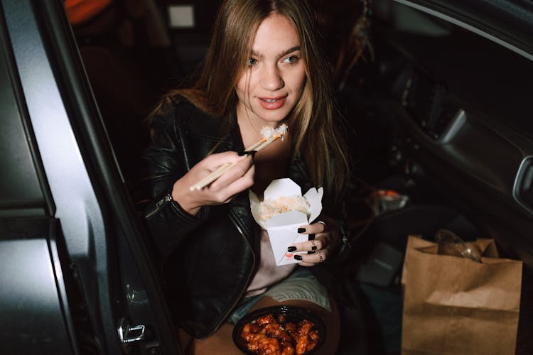 A Woman In Black Leather Jacket Sitting In The Car While Eating Foods