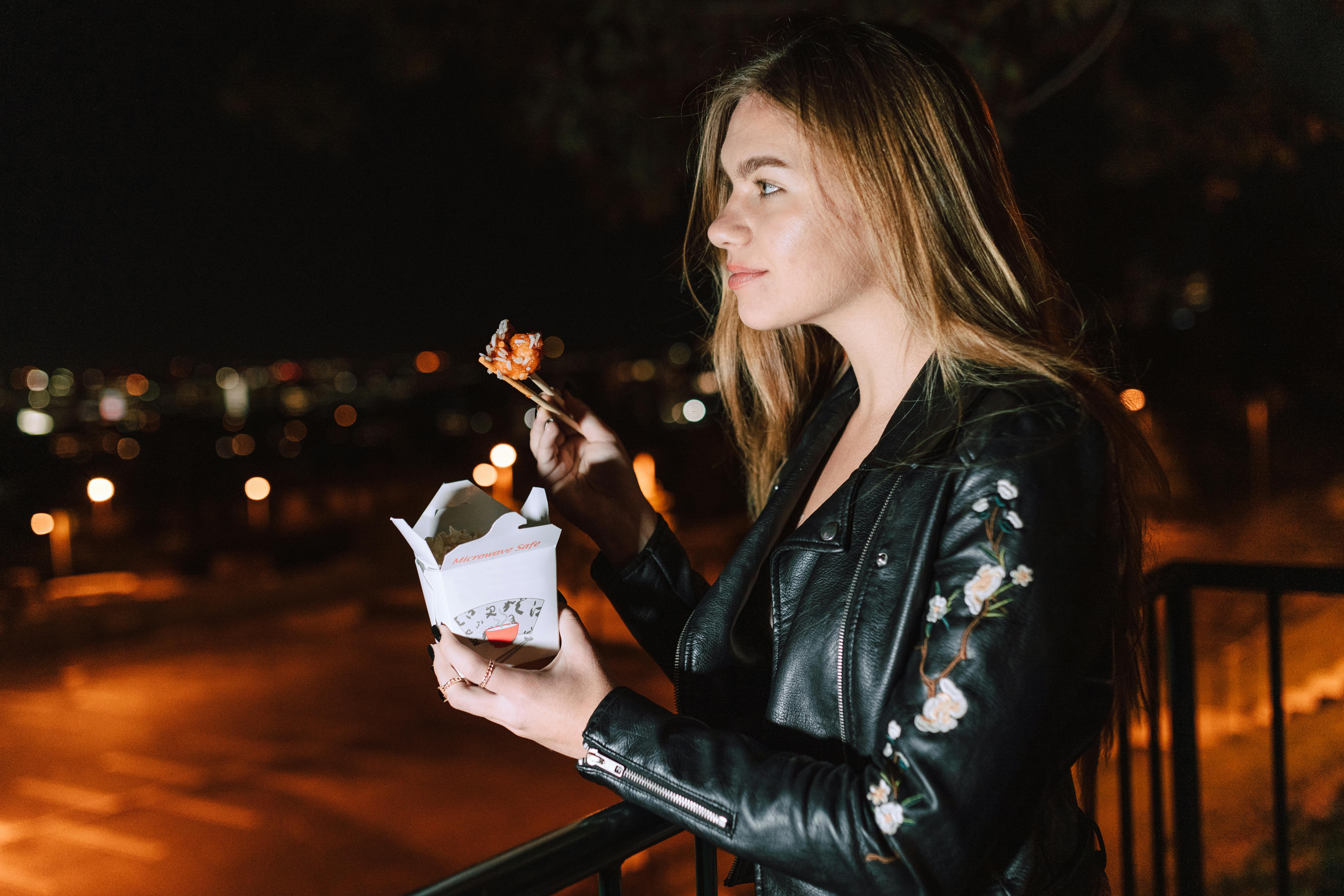 Young woman eating Asian takeout at night, city lights glowing in the background.
