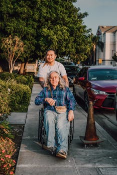 A caring scene of a young man pushing a senior in a wheelchair along a sidewalk.