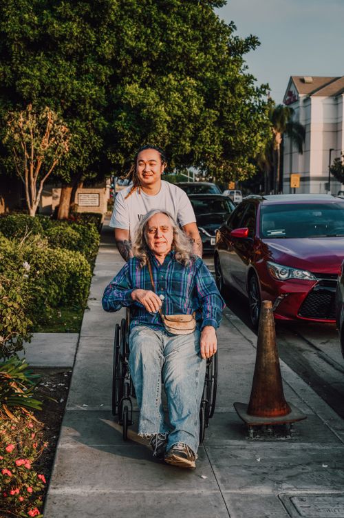 Free A caring scene of a young man pushing a senior in a wheelchair along a sidewalk. Stock Photo