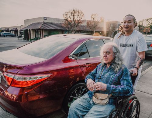 Free A volunteer helps an elderly man in a wheelchair near parked cars on a sunny day. Stock Photo