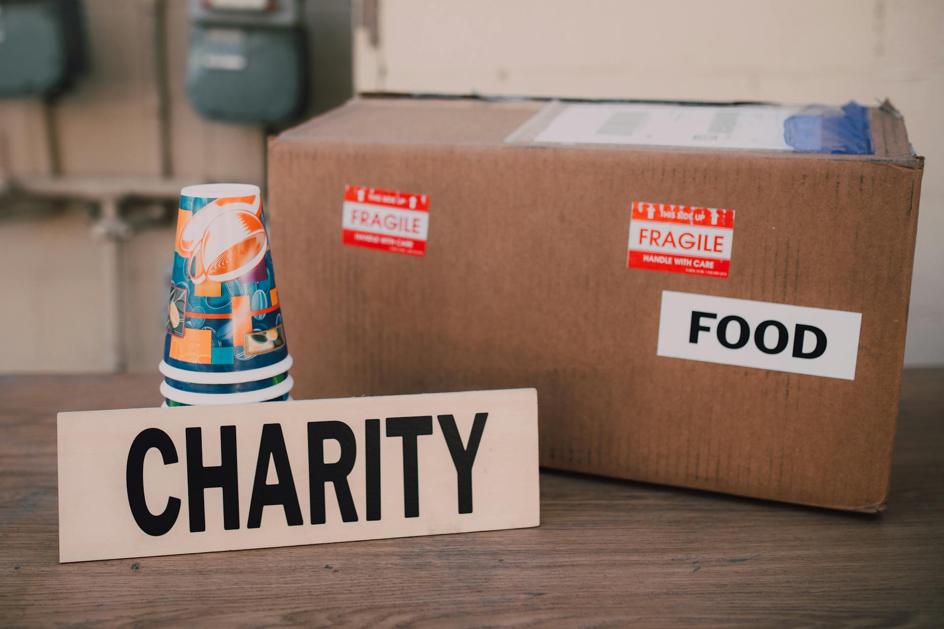 A Cardboard Box with Food Label Beside a Charity Sign and Paper Cups