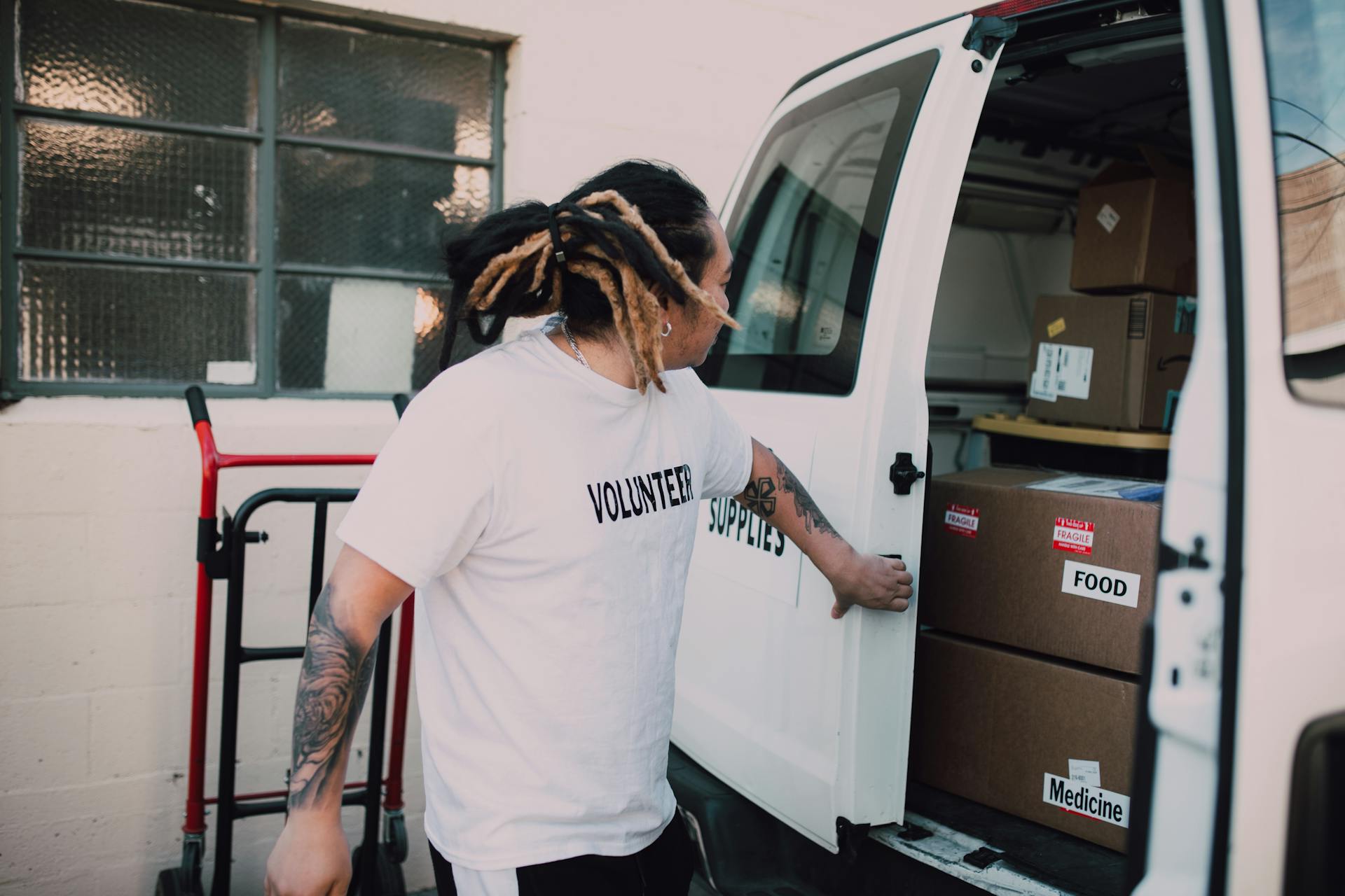 A Man Wearing Volunteer Shirt Standing Beside a Van with Boxes of Food and Medicine