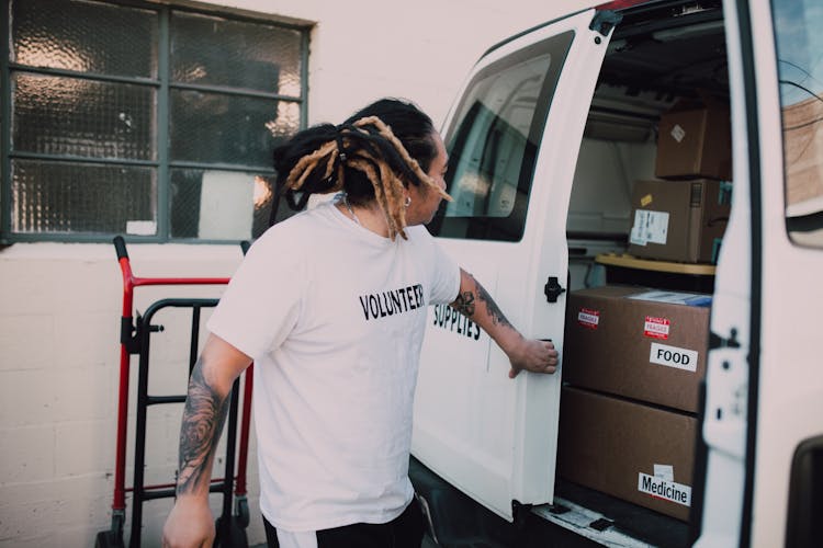 A Man Wearing Volunteer Shirt Standing Beside A Van With Boxes Of Food And Medicine