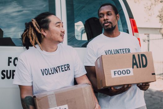 Two volunteers wearing white shirts distribute food aid boxes outdoors as part of a charity effort.