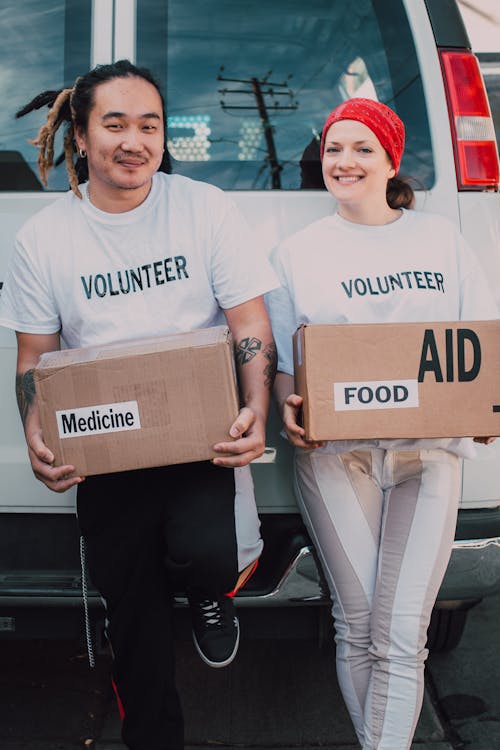 Free Man and Woman Carrying Medicine and Food Labelled Cardboard Boxes Behind a White Van Stock Photo