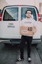 Man Wearing White Crew Neck Volunteer T-shirt Holding a Food Labelled Cardboard Box Behind White Van