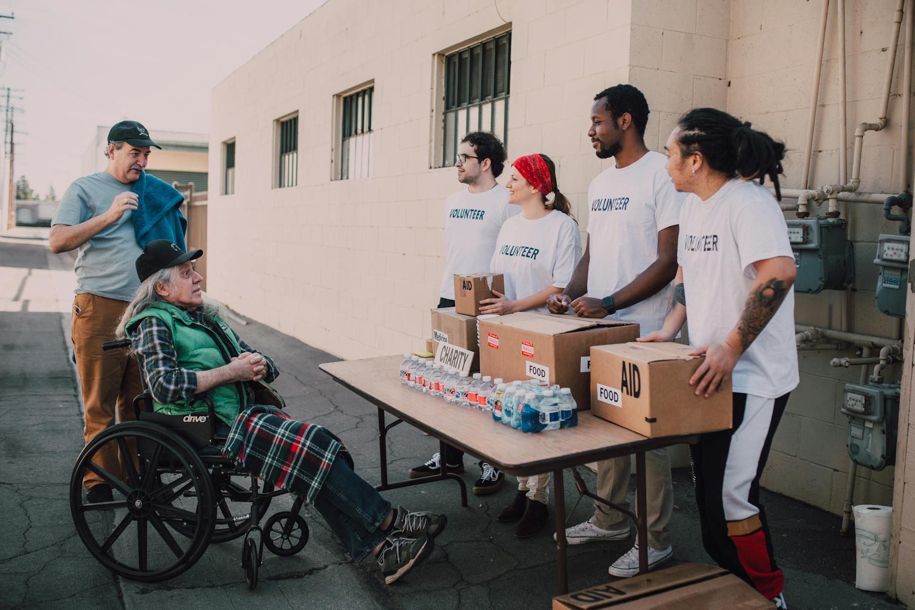 Volunteers assist a senior adult in a wheelchair by distributing aid packages outdoors.