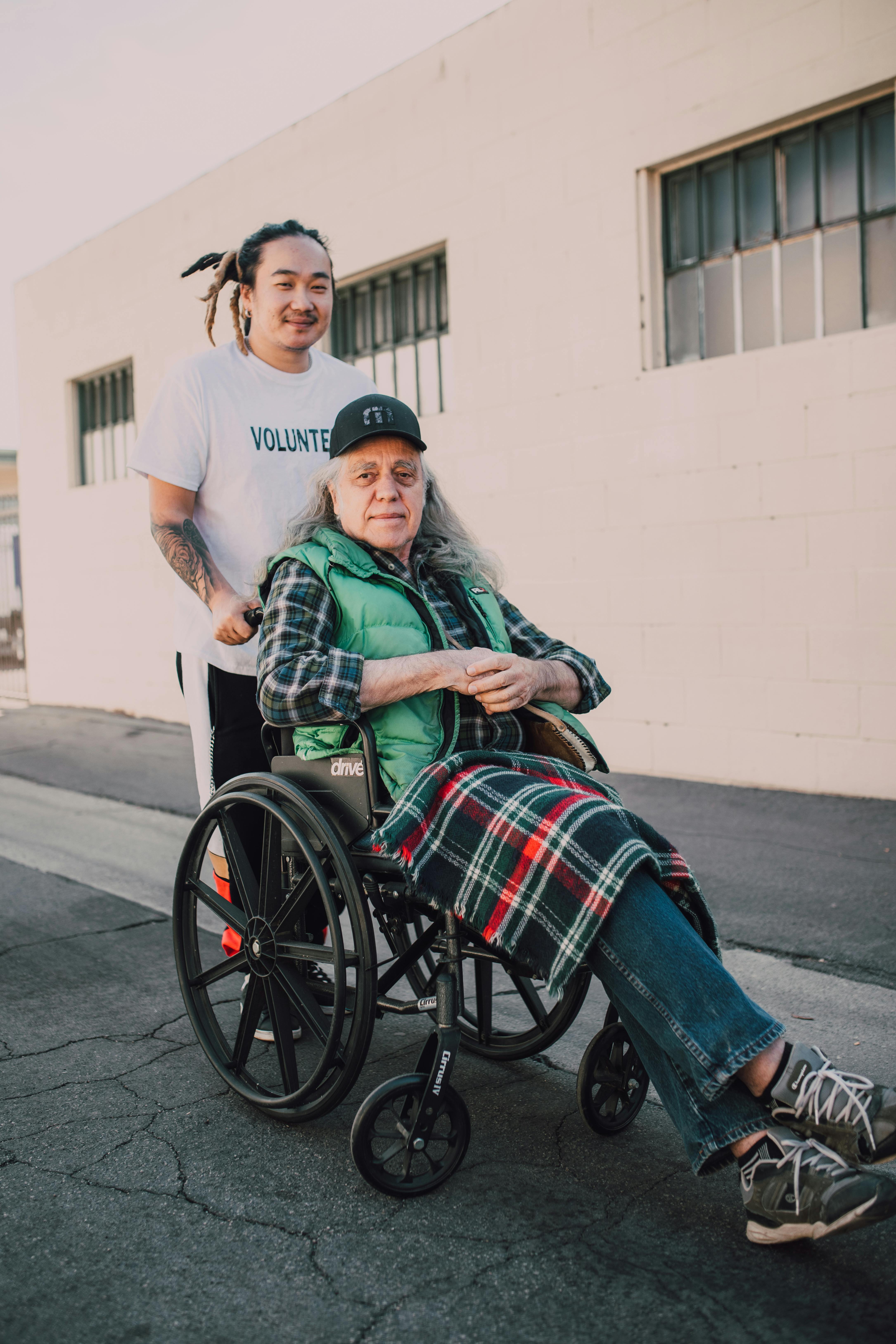 A young volunteer helps an elderly man in a wheelchair on a sunny street.
