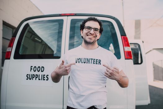 Happy volunteer showing off t-shirt standing in front of a food supplies delivery van outdoors.