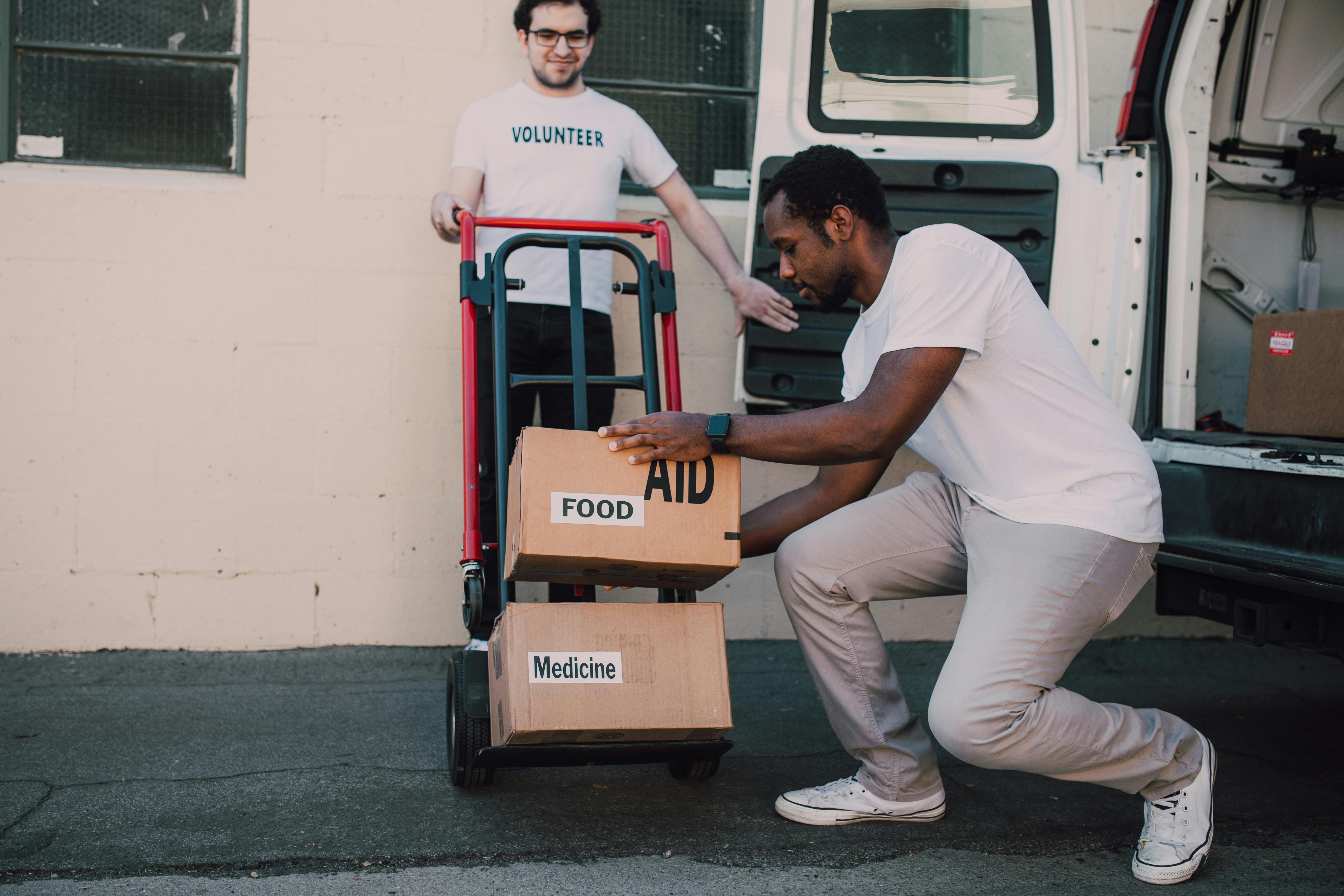 Two volunteers load boxes labelled food aid and medicine onto a trolley beside a van.