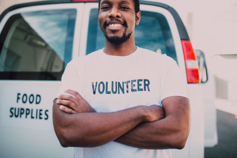 A smiling Black man in a white VOLUNTEER T-shirt stands with his arms crossed in front of a white outreach van marked FOOD SUPPLIES, the kind of community vehicle that brings supplies and care to people where they are.