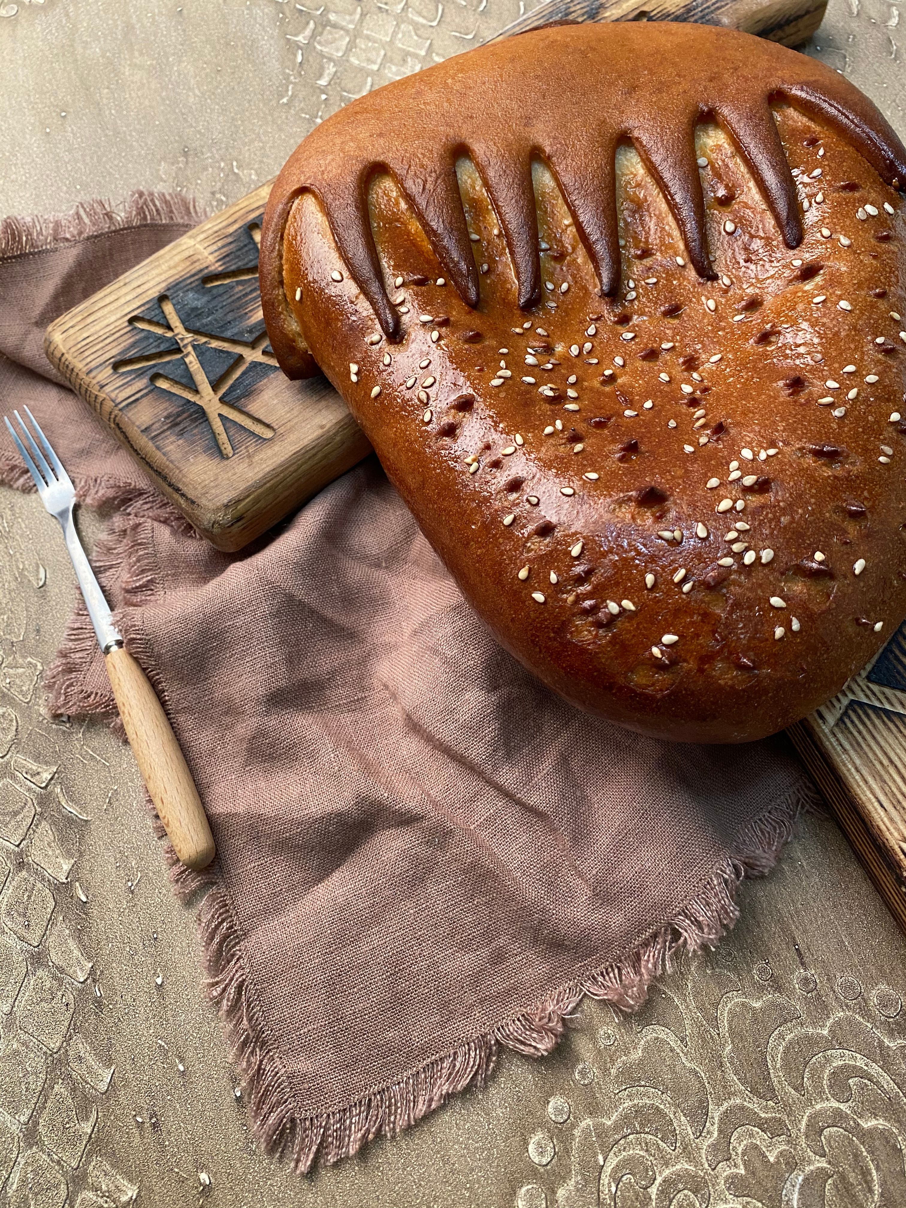 Freshly baked bread loaf with sesame seeds on rustic cutting boards and a fork on a beige cloth.