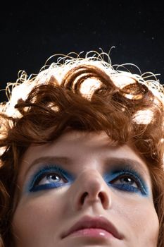 Close-up of a young woman with curly hair and striking blue eyeshadow, captured in a compelling upward gaze.