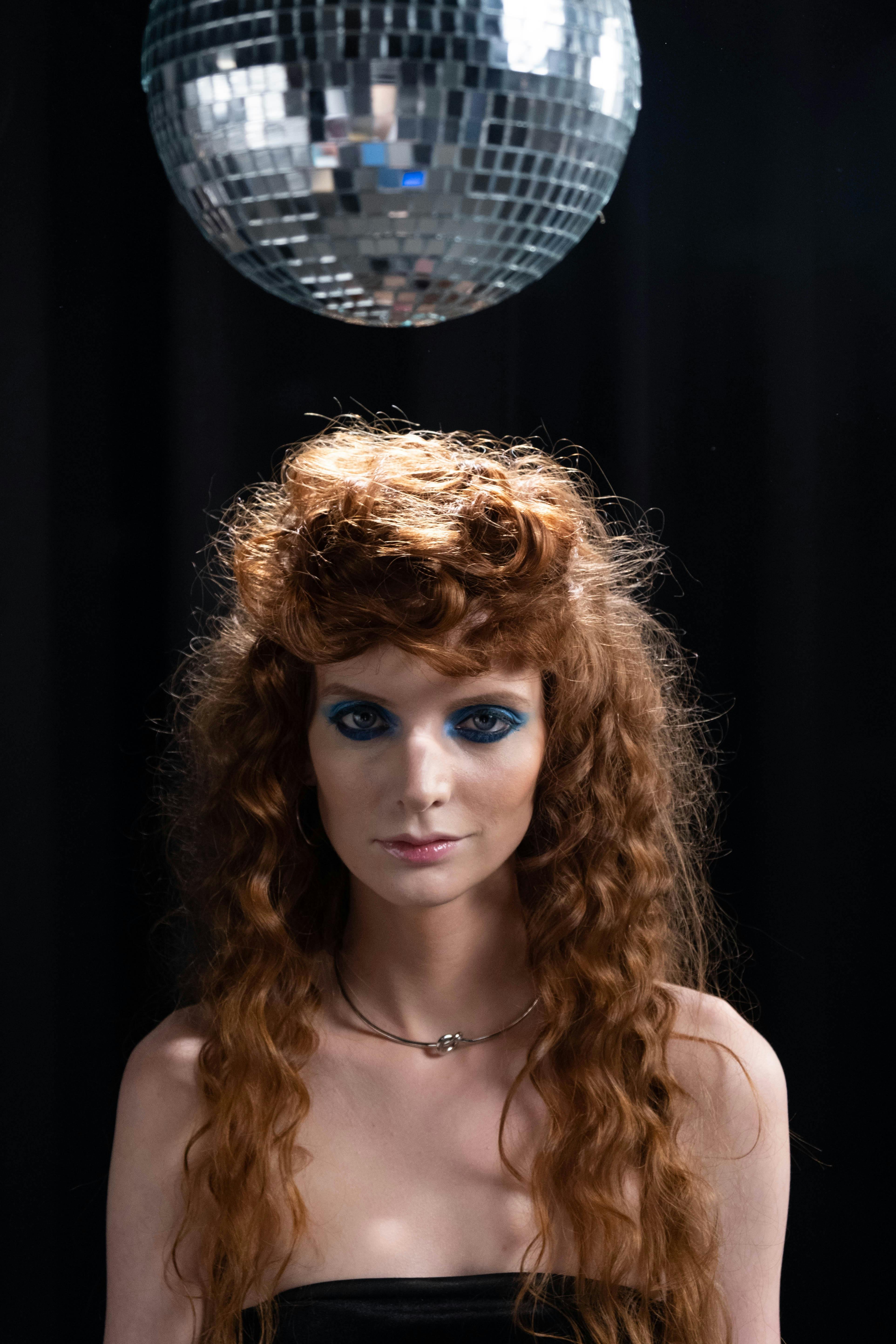A young woman with bold blue eyeshadow stands under a disco ball indoors.