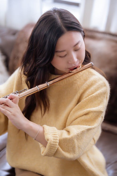 Young Asian woman in casual clothes practicing melody on flute while sitting on couch in bright room