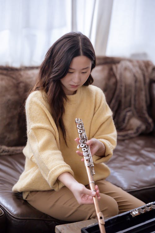 Serious young ethnic female in casual outfit looking at flute while sitting on brown sofa near curtains and table with case in light apartment