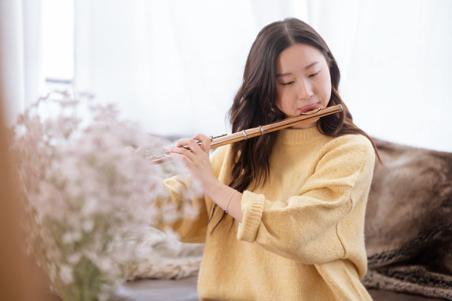 Focused Asian female musician playing melody on flute while sitting in light room with blooming flowers at home during rehearsal