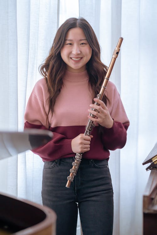 Happy young ethnic woman in casual outfit with flute in hands standing near wooden piano in light room while looking at camera near curtains