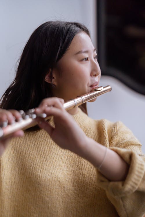 Thoughtful young Asian lady in warm sweater playing on flute in light classroom