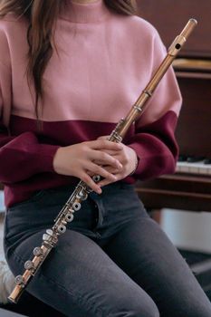 Crop anonymous woman in warm sweater and jeans with flute sitting on stool near piano in bright room