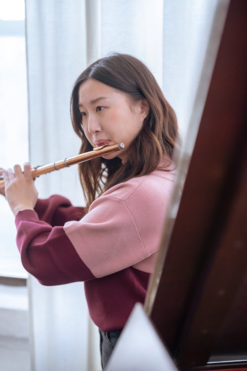 Pensive young Asian lady in warm sweater playing on flute in light classroom near window with curtains