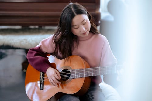 Thoughtful young Asian lady in casual clothes sitting on pouf while playing on guitar in bright classroom